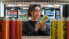 A woman in a black sweatshirt stands in a beverage store refrigerator, holding and reading a yellow can of MYND energy drink. Colorful energy drink cans are blurred in the foreground and background.
