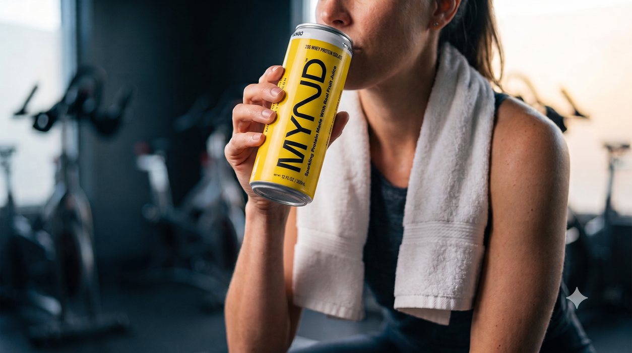A woman in a gym setting drinking from a yellow can of MYND energy drink, with a white towel draped over her shoulders.
