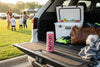 A pink MYND energy drink can stands on the open tailgate of a white SUV, with a white YETI cooler, brown leather bag, neon green sneakers, and a soccer ball nearby. The scene is set outdoors at a park or field with people blurred in the background during golden hour.